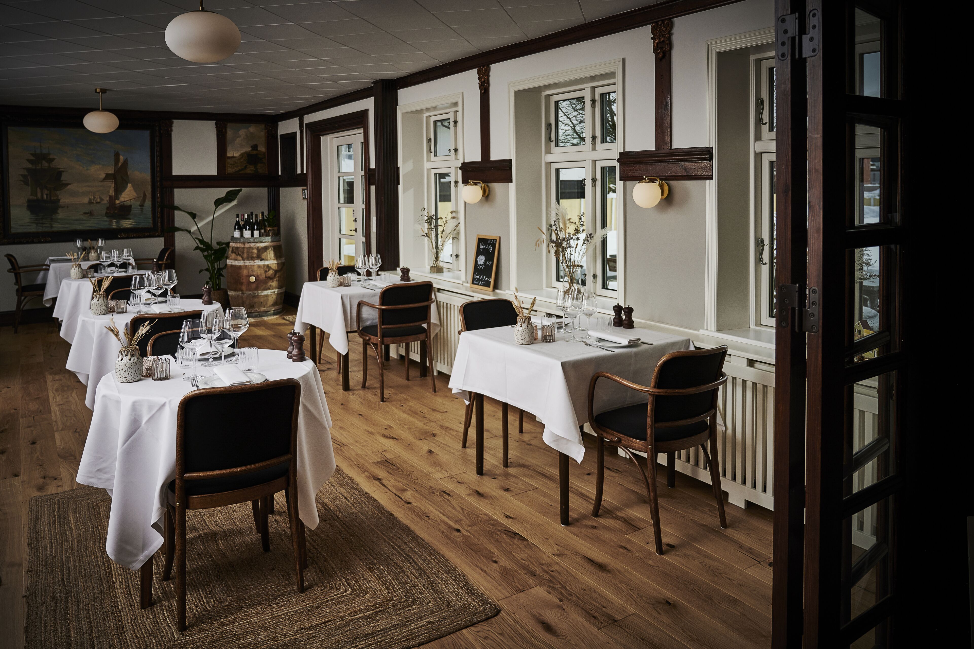 A dining area with multiple tables set for guests, wooden floors, and large windows with views.