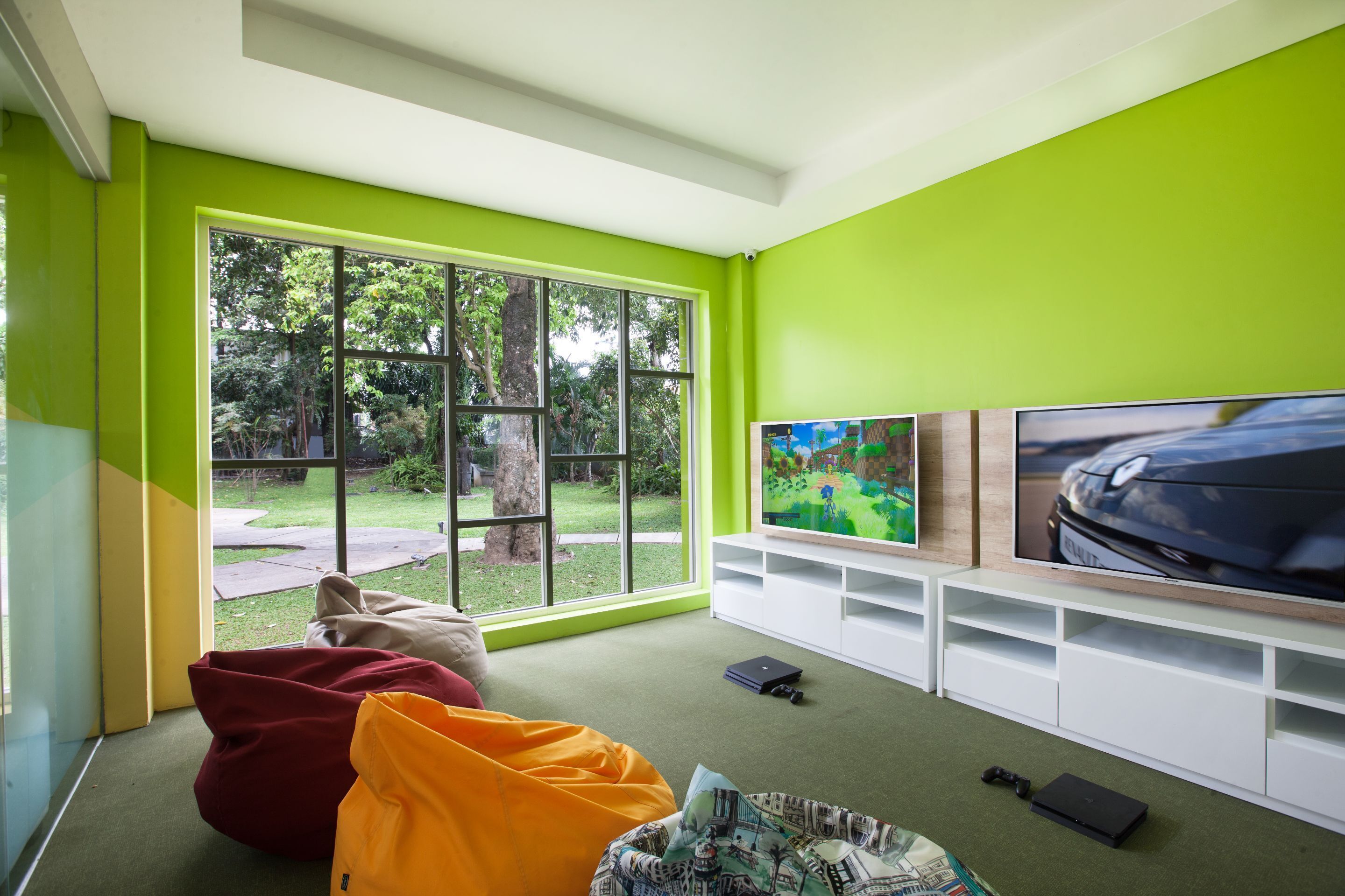 Una sala de estar moderna con un ventanal grande, paredes verdes, un televisor de pantalla plana y una consola de videojuegos.