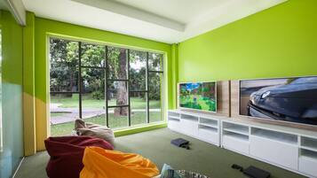Una sala de estar moderna con un ventanal grande, paredes verdes, un televisor de pantalla plana y una consola de videojuegos.