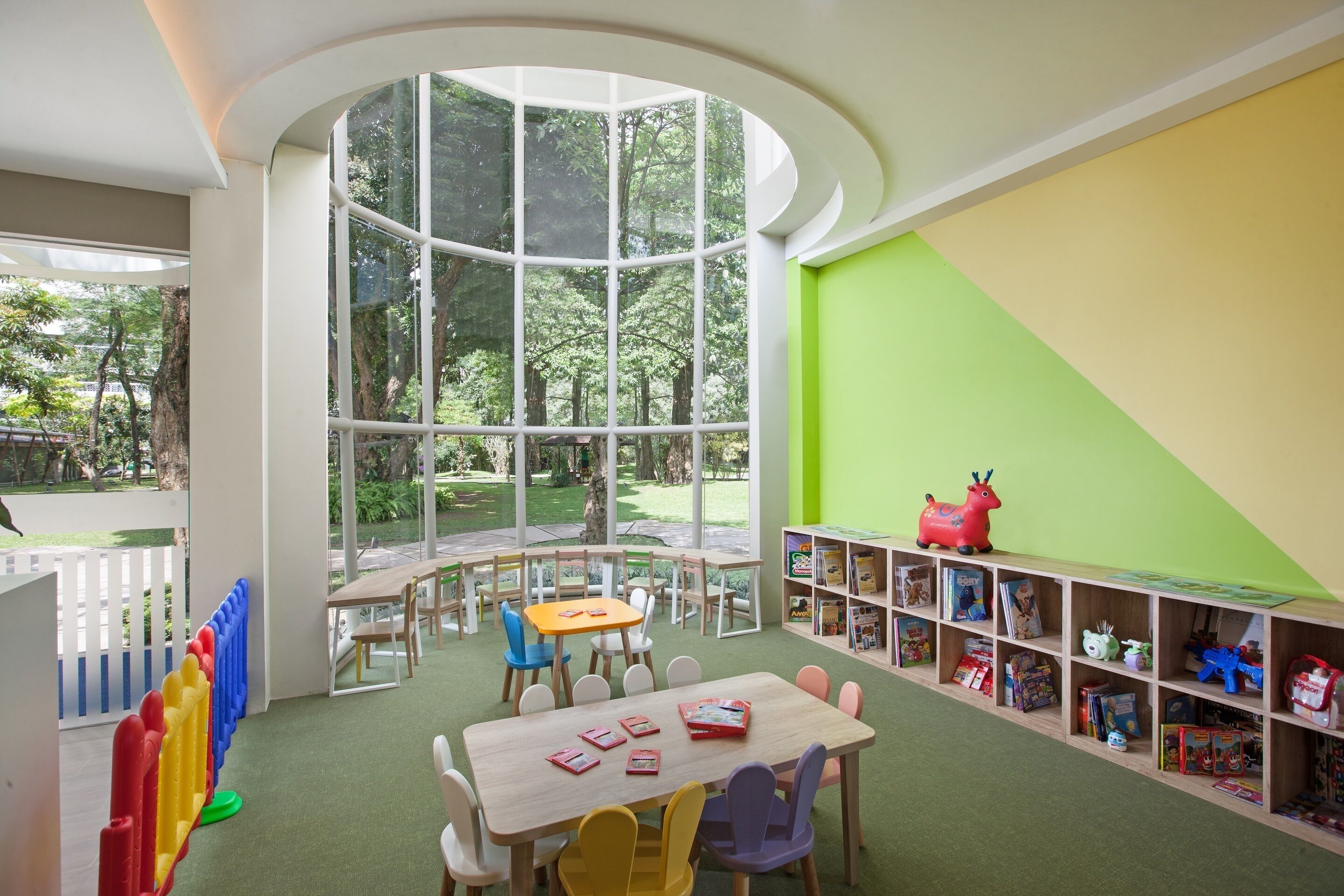 Una sala giochi per bambini con un'ampia finestra, un tavolo di legno, sedie e una libreria piena di giochi.