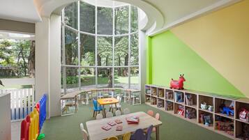 Una sala giochi per bambini con un'ampia finestra, un tavolo di legno, sedie e una libreria piena di giochi.