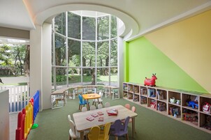 Una sala giochi per bambini con un'ampia finestra, un tavolo di legno, sedie e una libreria piena di giochi.