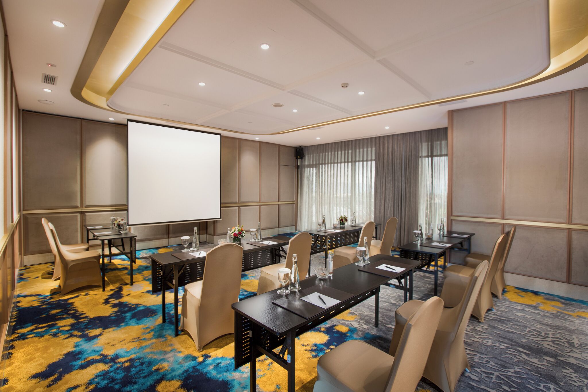 A conference room with a large screen, multiple tables, and chairs, carpeted floor, and a modern ceiling design.