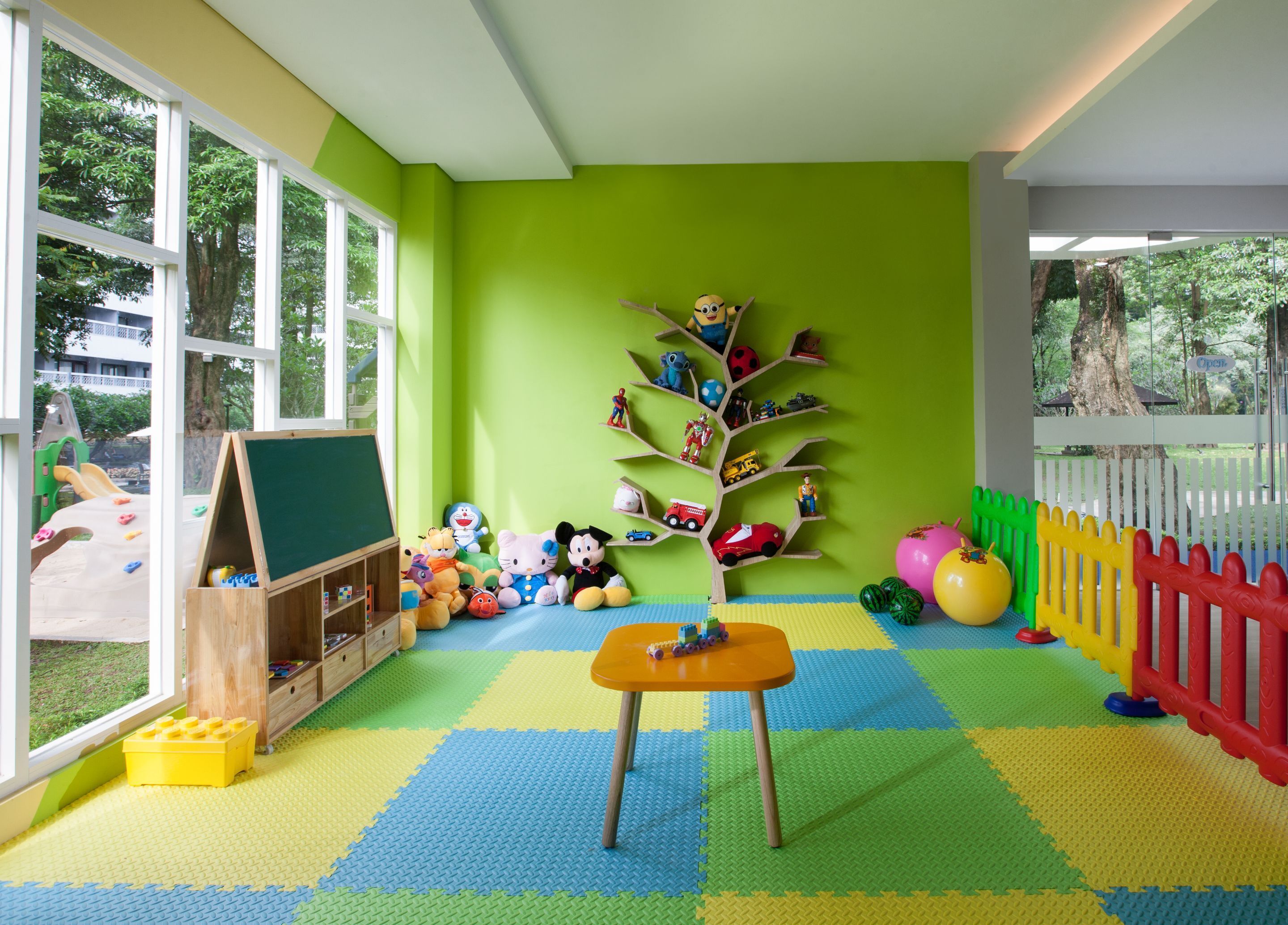 A brightly colored playroom with a large window, a wooden play table, and a toy tree shelf.