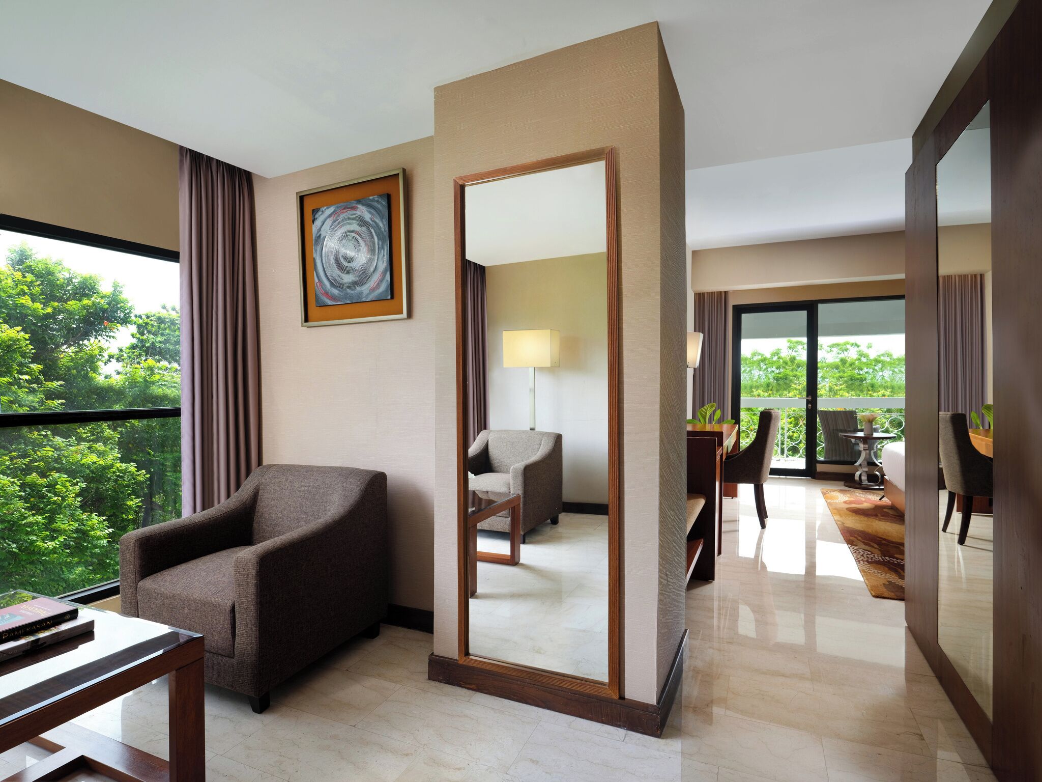 A hotel room with a large mirror, a chair, a table, and a view of greenery outside.