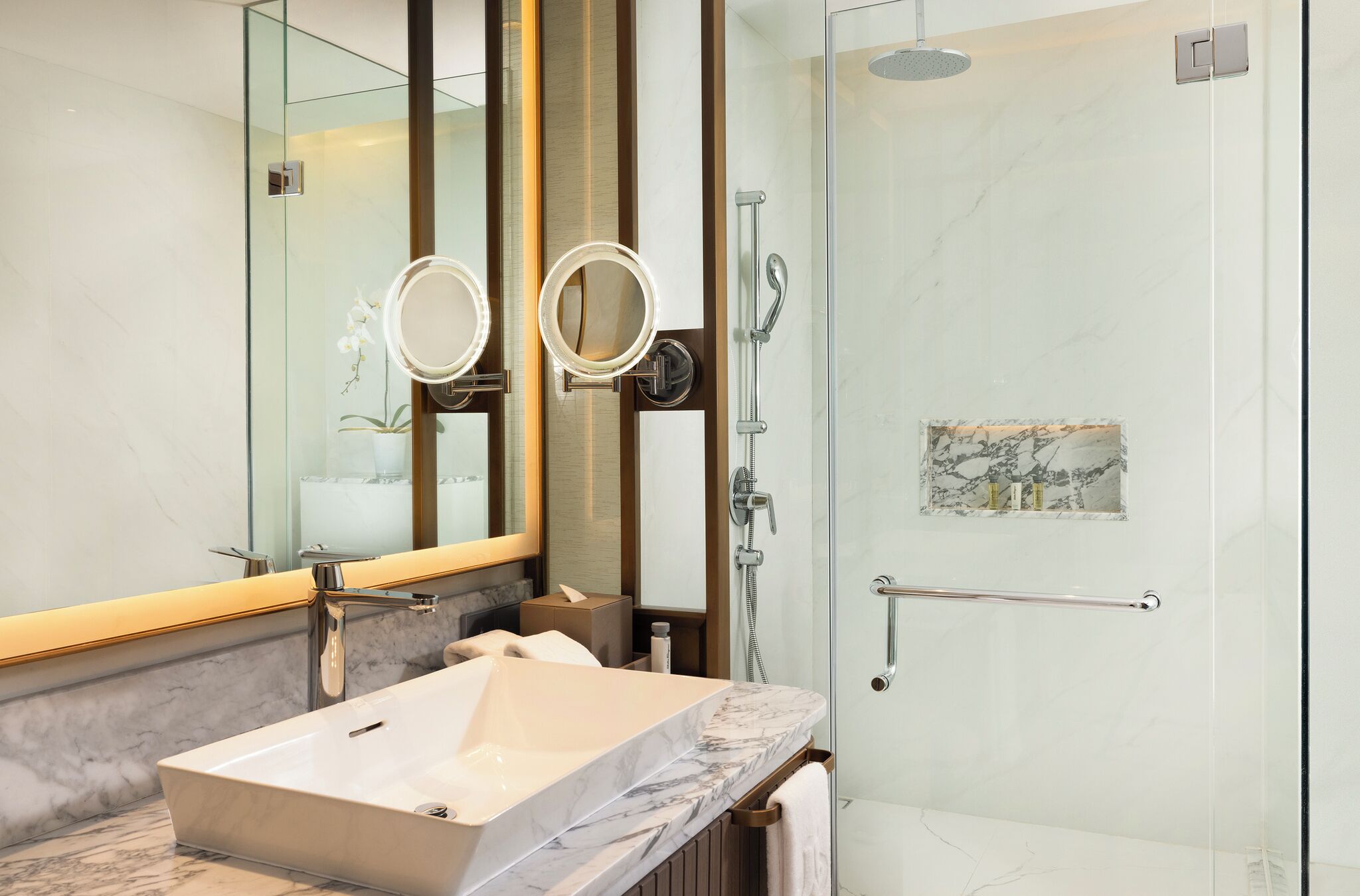 A modern bathroom with a marble sink, two round mirrors, a glass shower enclosure, and a towel rack.