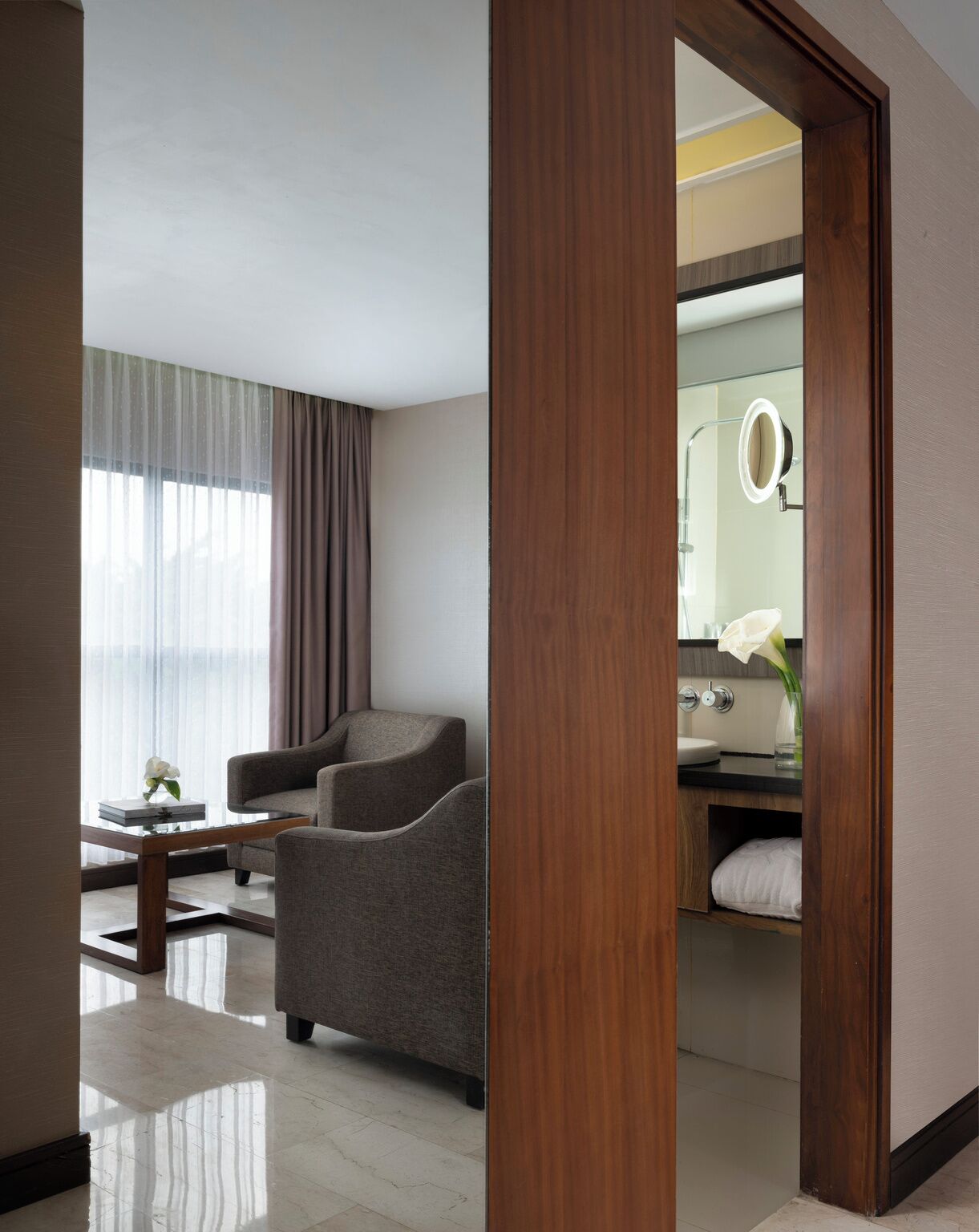 Ein modernes Hotelzimmer mit grauem Sessel, einem kleinen Tisch mit Buch und Blumen, einem Badezimmer mit Spiegel und Waschbecken sowie Blick durch eine Holztür.