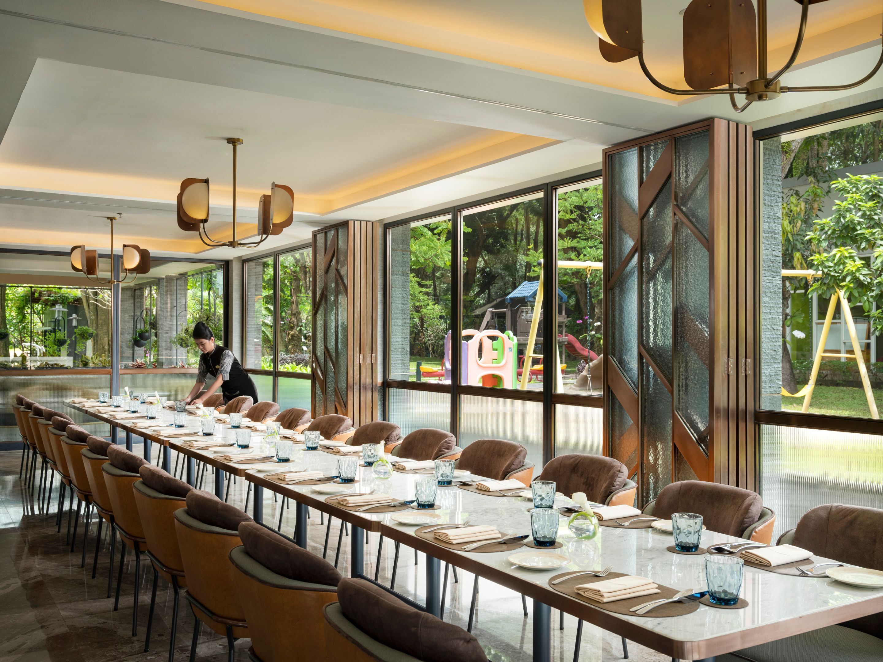 Una larga mesa de comedor lista para una comida, con sillas y una persona atendiendo a ella.
