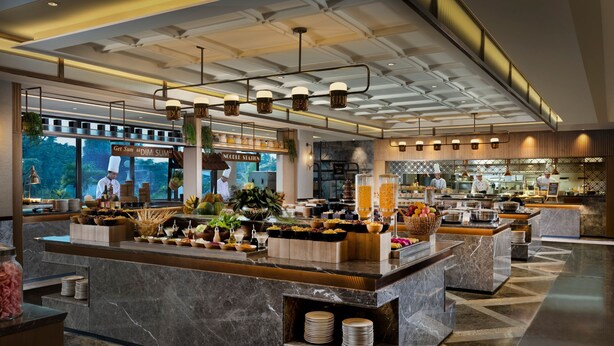A modern hotel lobby with a marble reception desk, a large window with a view of greenery, and a visible restaurant area in the background.