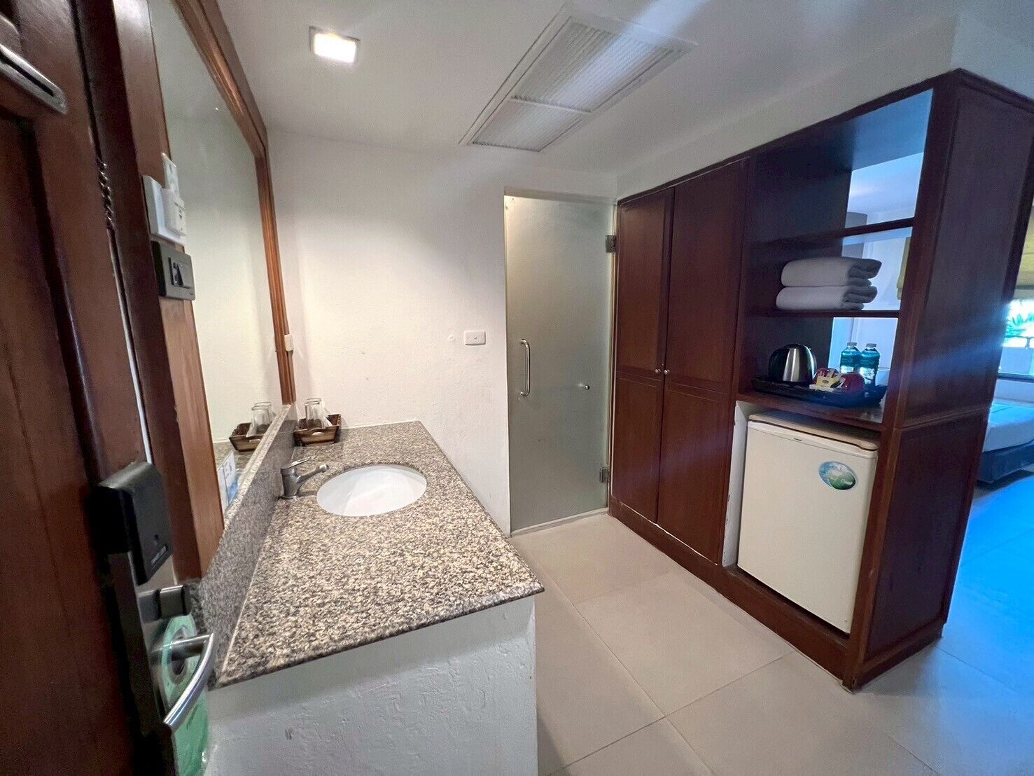 A bathroom with a granite countertop sink, a mirror, a cabinet with towels, and a small refrigerator.