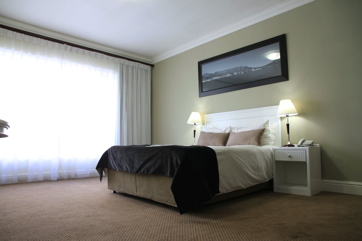 Una habitación de hotel con una cama grande, dos mesitas de noche con lámparas, un cuadro en la pared y vistas al exterior a través de cortinas traslúcidas.