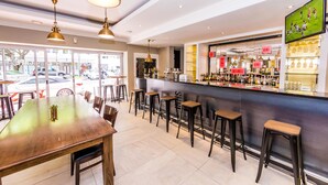 A bar area with a long wooden table and multiple stools, a counter with a TV screen, and a view of the street outside. - The New Tulbagh Hotel (Cape Town)