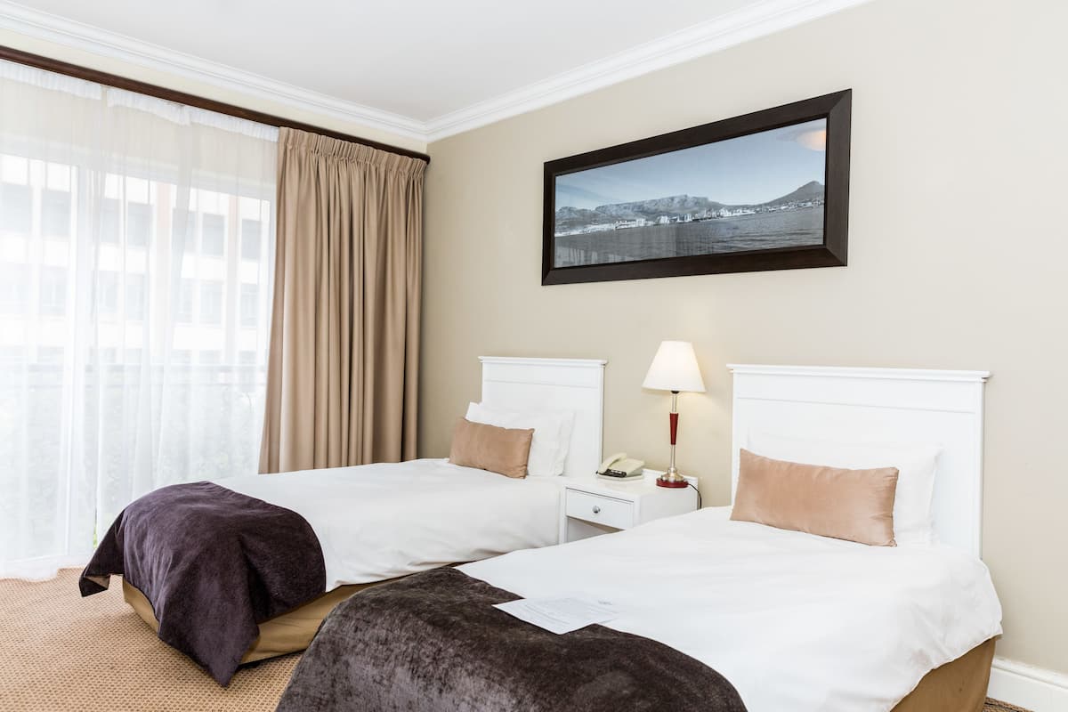 Habitación de hotel con dos camas, una mesita de noche con lámpara, un cuadro en la pared y una ventana con cortinas.