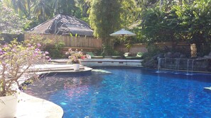 A backyard with a swimming pool, a wooden deck, a thatched-roof structure, and a waterfall feature. - The Watergarden (Karangasem)