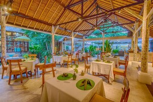 Un área de comedor al aire libre con mesas y sillas, rodeada de abundante follaje y una estructura de techo de paja.