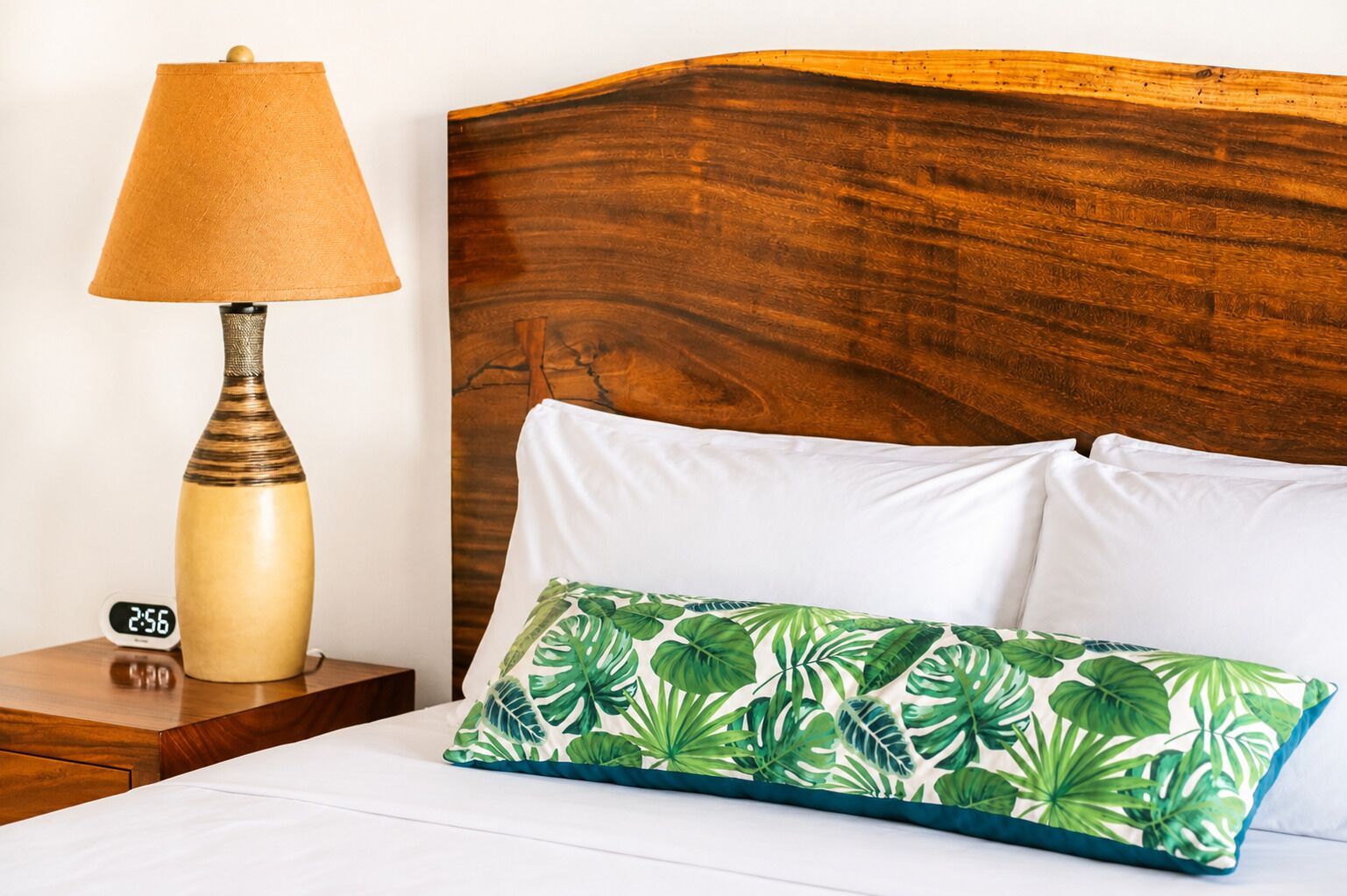 A bed with white pillows and a green leaf-patterned pillow, a wooden nightstand with a lamp, and a digital clock.