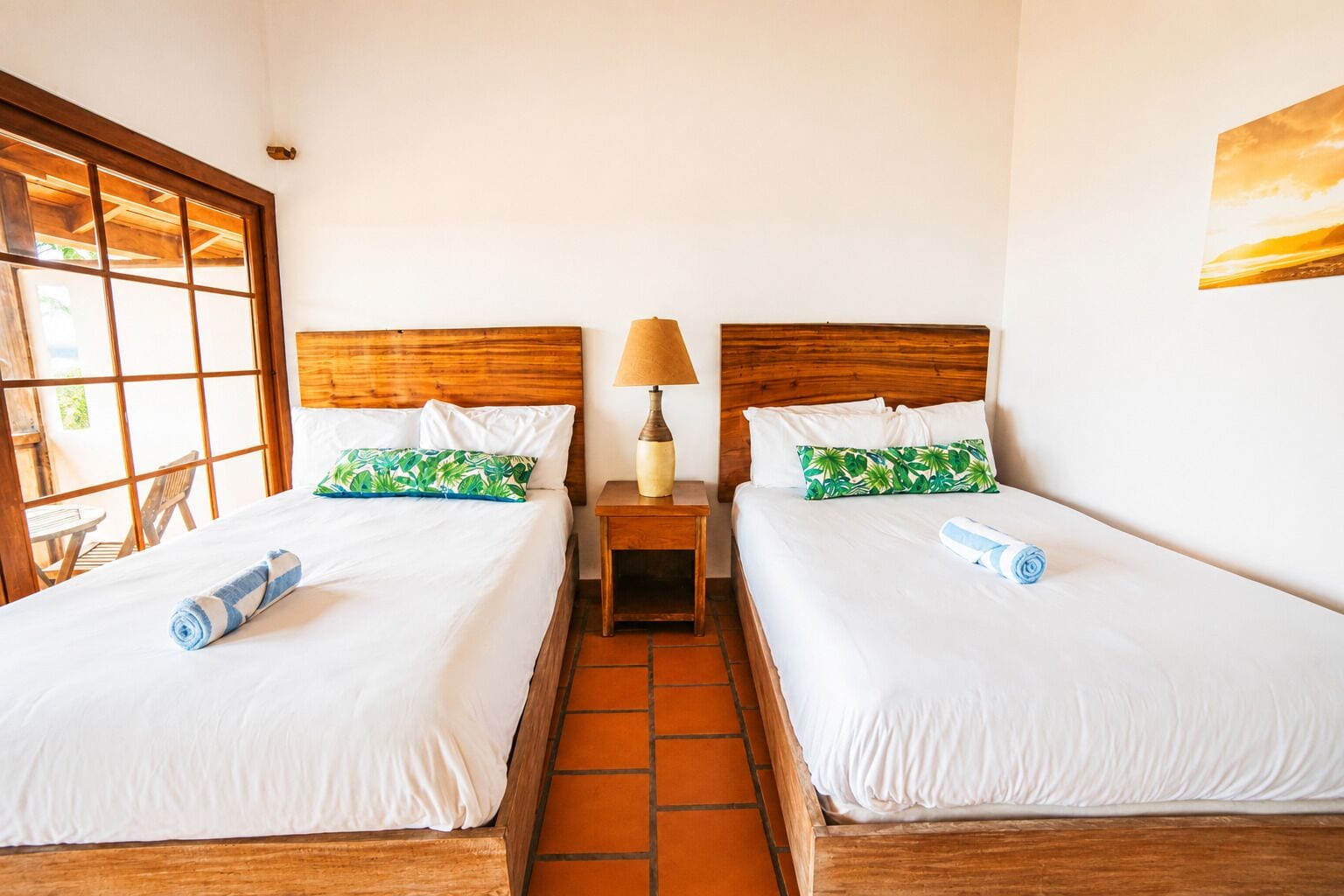 A hotel room with two beds, a nightstand with a lamp, and a window with a view.