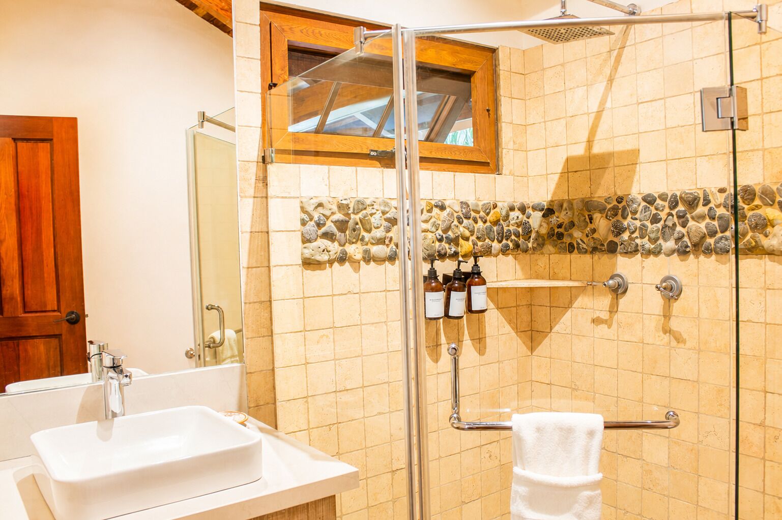 Baño con cabina de ducha de vidrio, lavamanos, espejo, toallero y ventana con marco de madera.