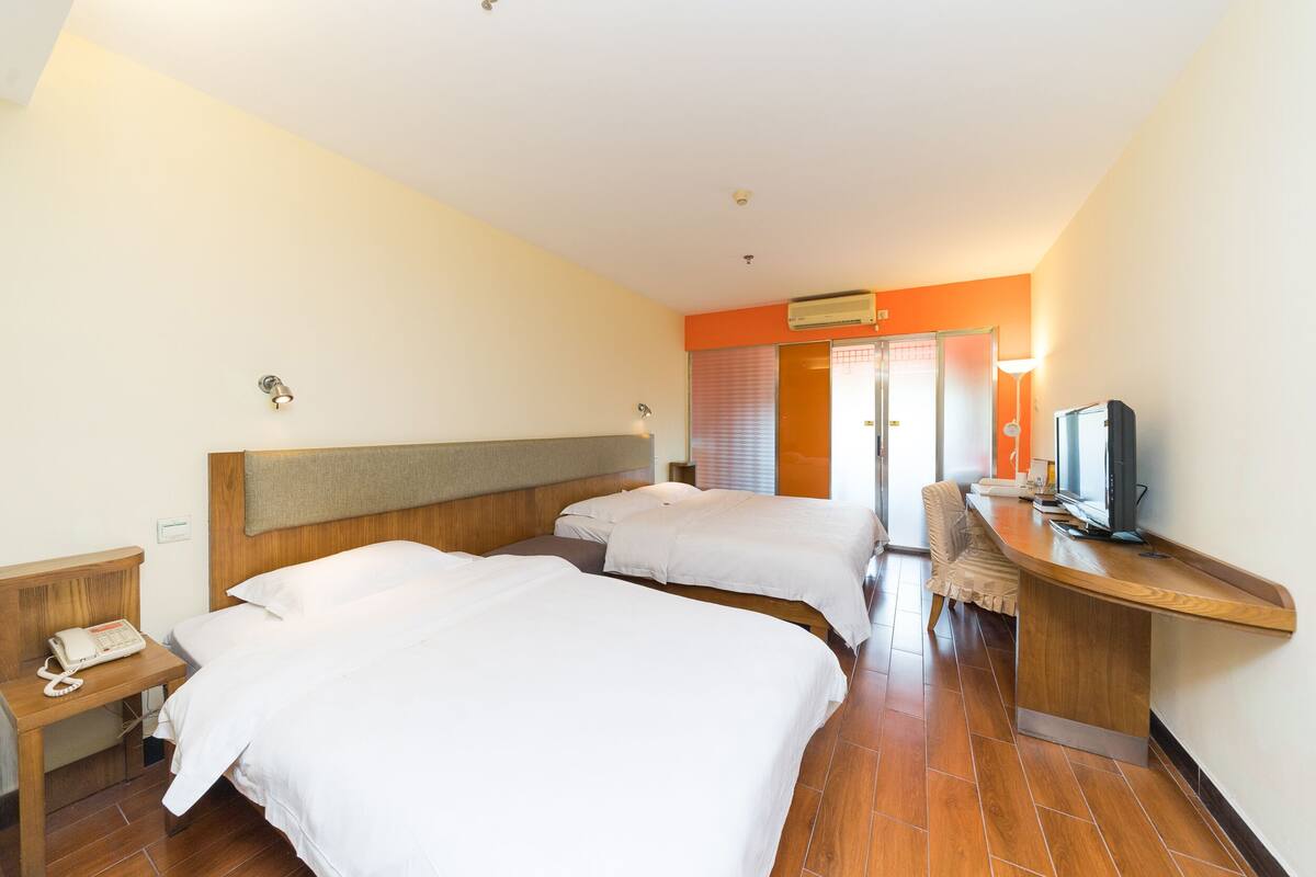 A hotel room with two beds, a desk with a computer, a telephone, and an air conditioning unit.