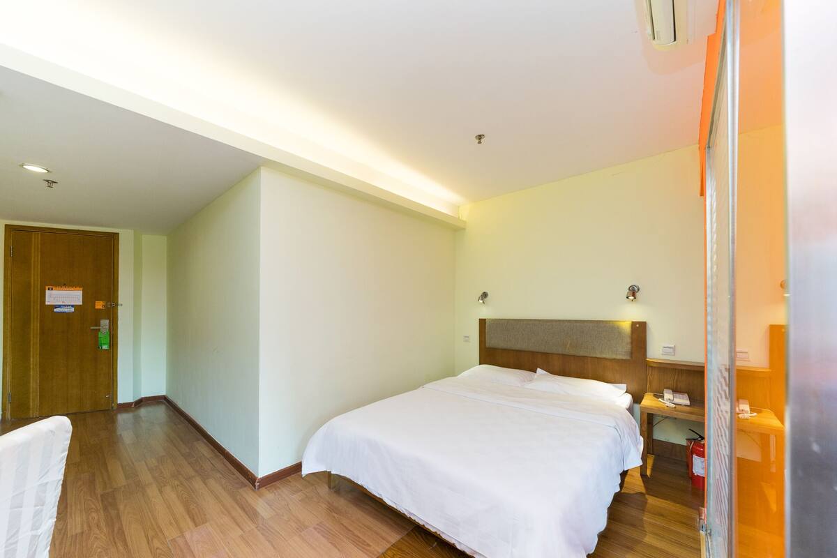 A hotel room with a large bed, a wooden headboard, a bedside table with a lamp, and a small desk with a chair.