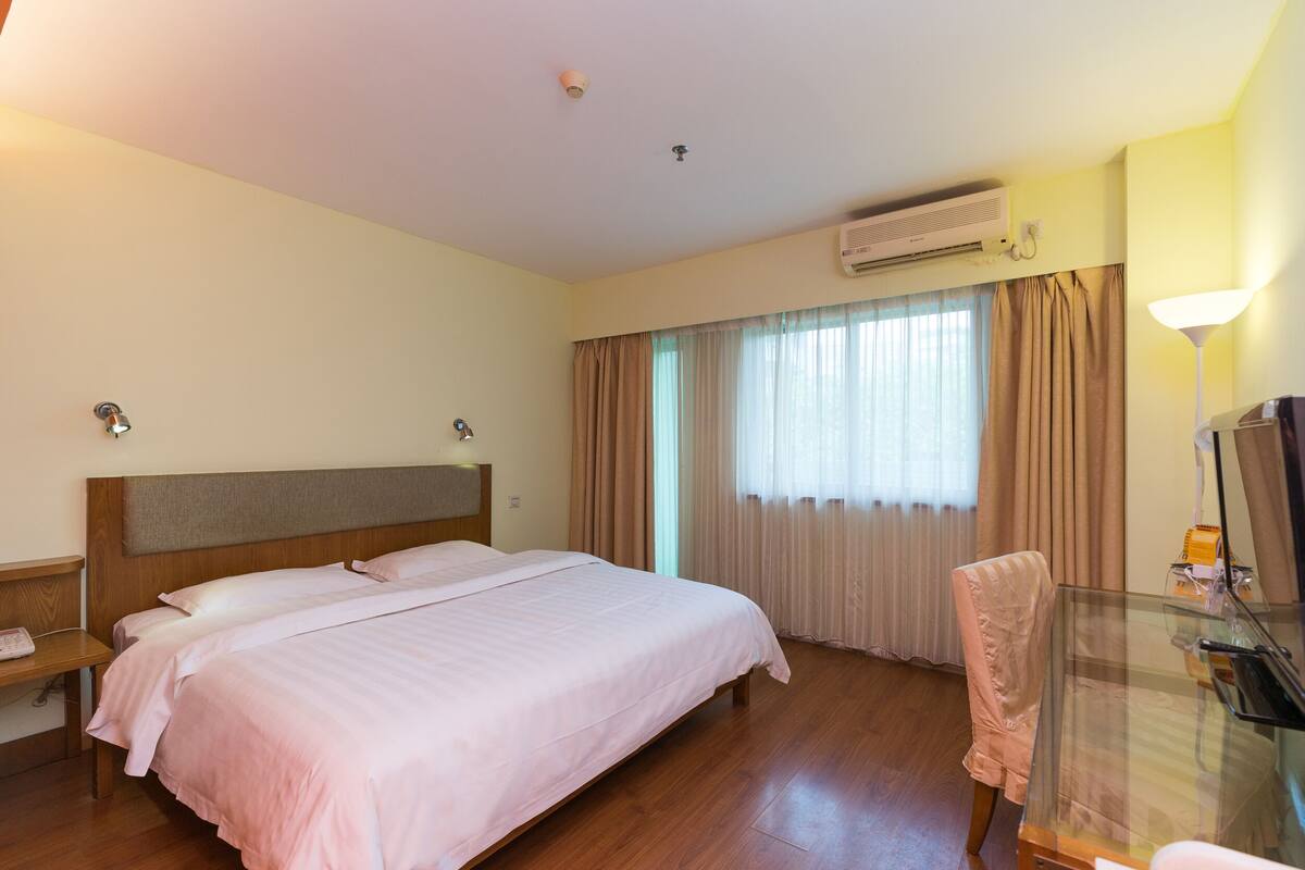A hotel room with a large bed, a desk with a chair, an air conditioning unit, and a window with curtains.