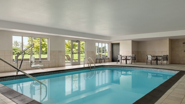 An indoor swimming pool with clear glass walls, a tiled floor, and a seating area with tables and chairs. - Holiday Inn Express Hotel & Suites NORTH FREMONT by IHG (Fremont)