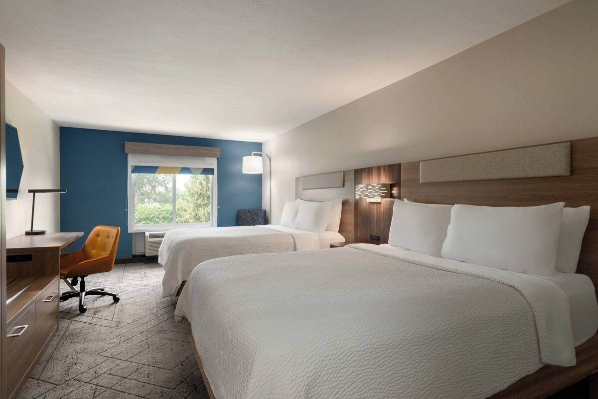 A hotel room with two beds, a desk, a chair, and a window with a view of greenery.