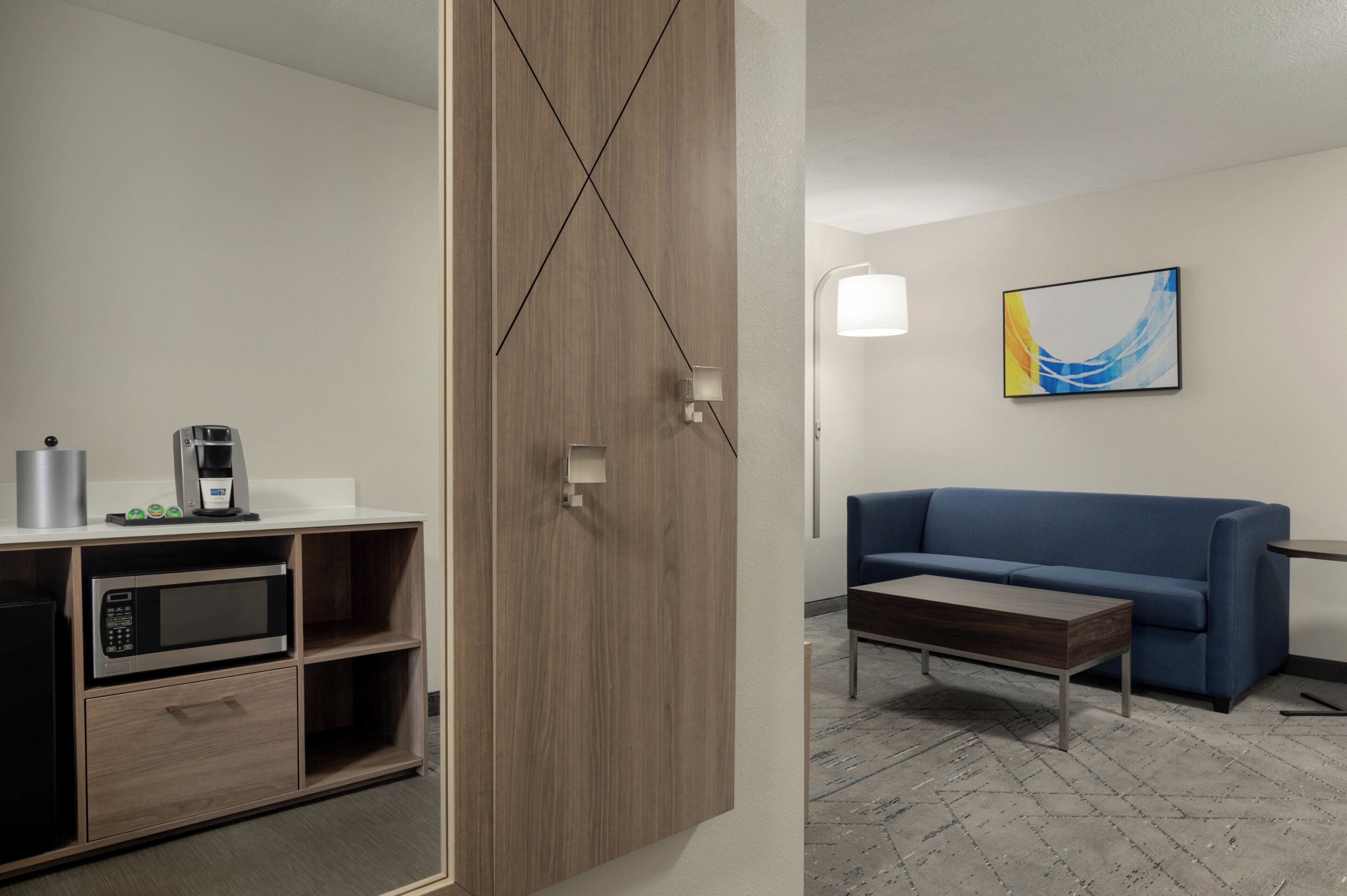 A modern hotel room with a kitchenette, a blue sofa, a coffee table, and a contemporary abstract painting.