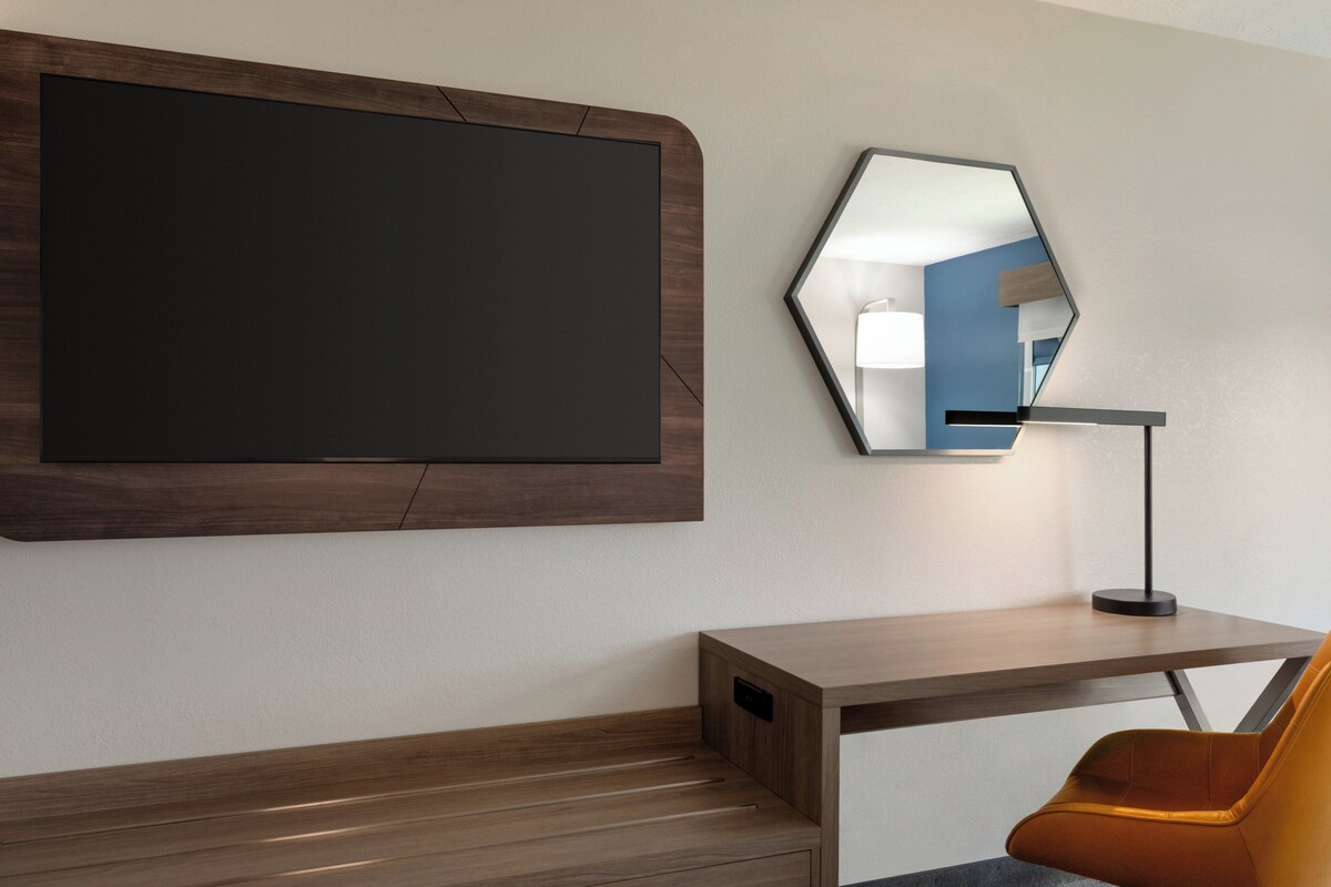 A modern hotel room with a large flat-screen TV mounted on the wall, a hexagonal mirror above a desk, and a single orange chair.