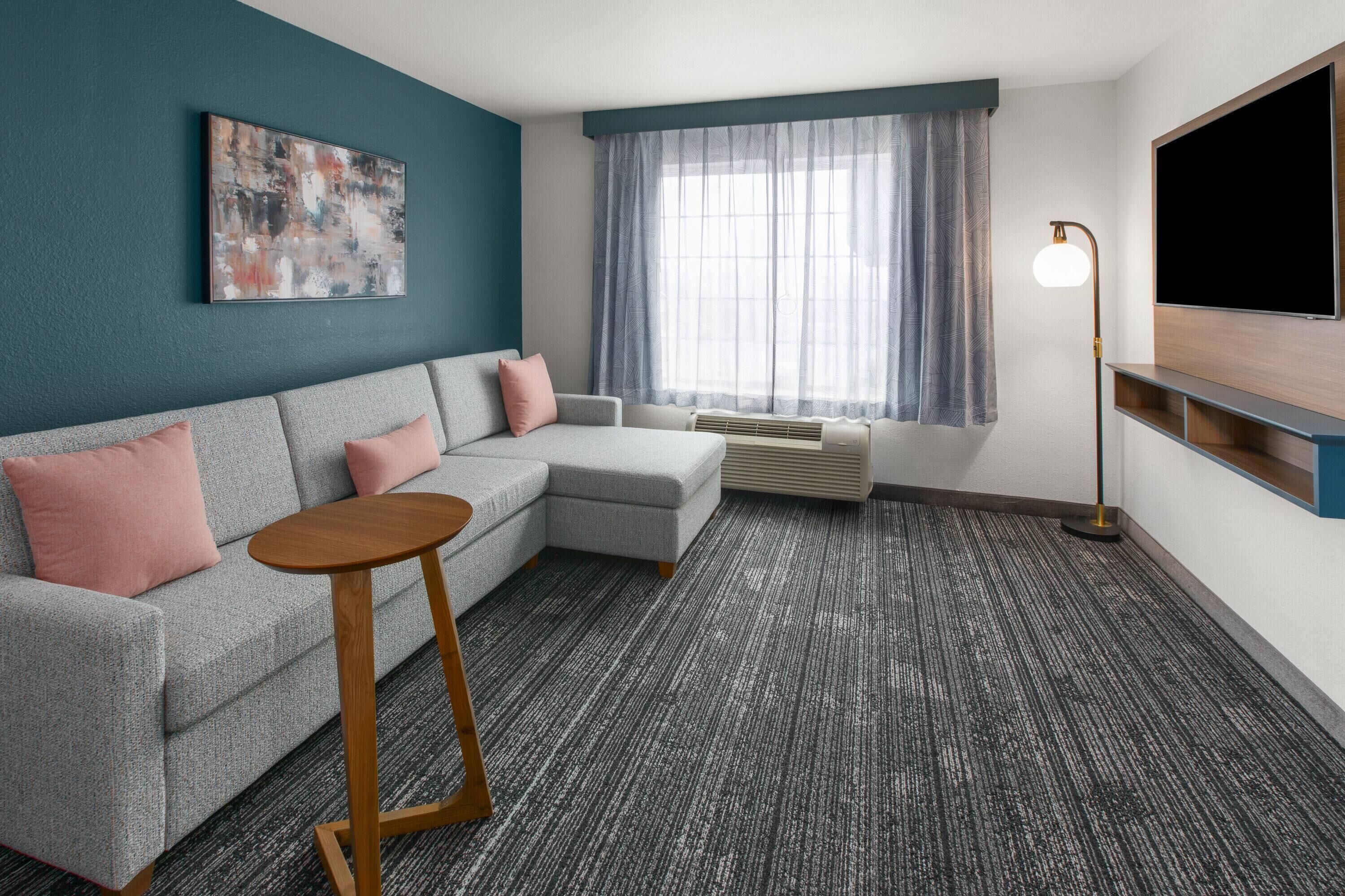 A modern living room with a grey sofa, a small round table, a large window with sheer curtains, a flat-screen TV, and a floor lamp.