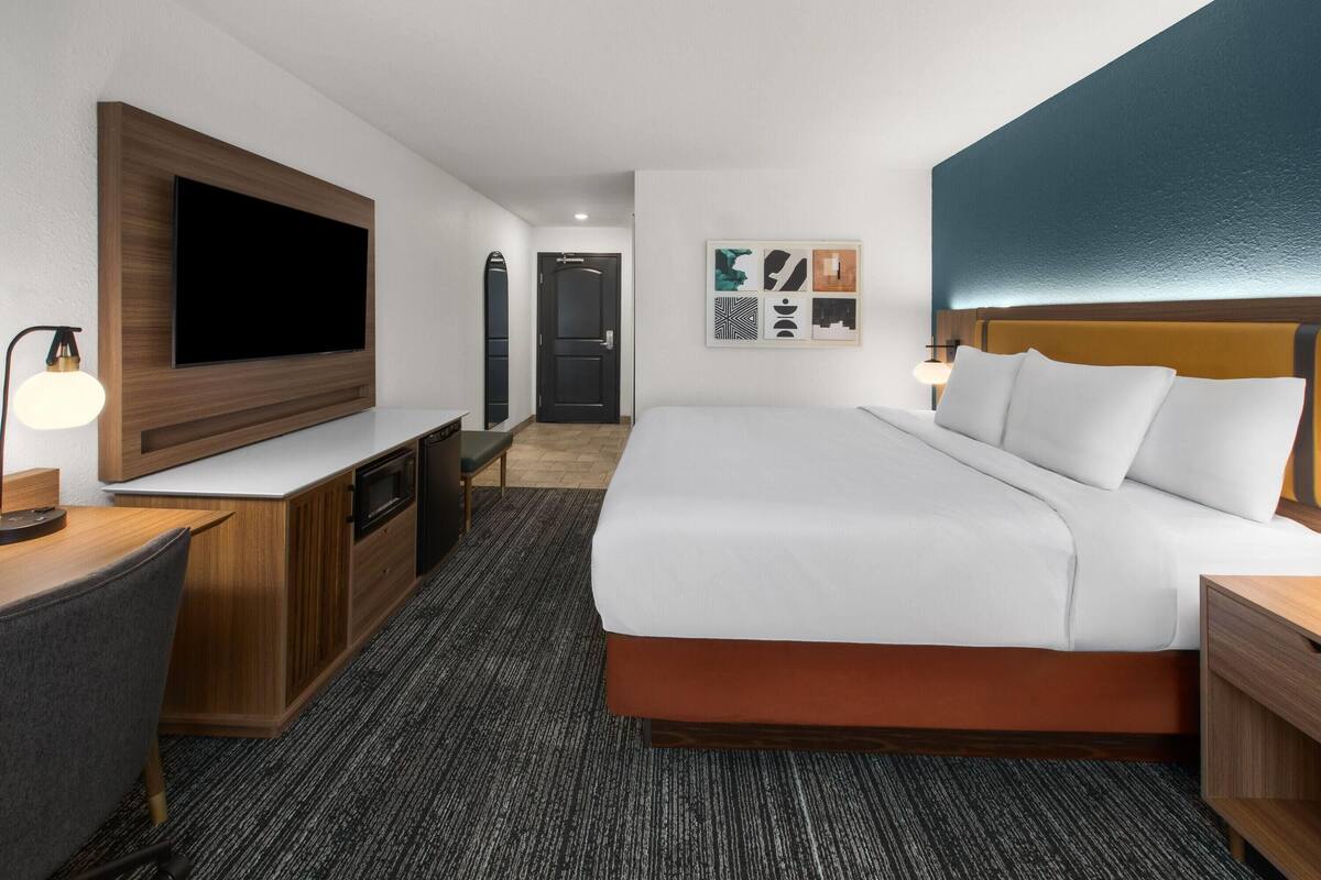 Una habitación de hotel con cama, escritorio, televisor, puerta y obras de arte en la pared.