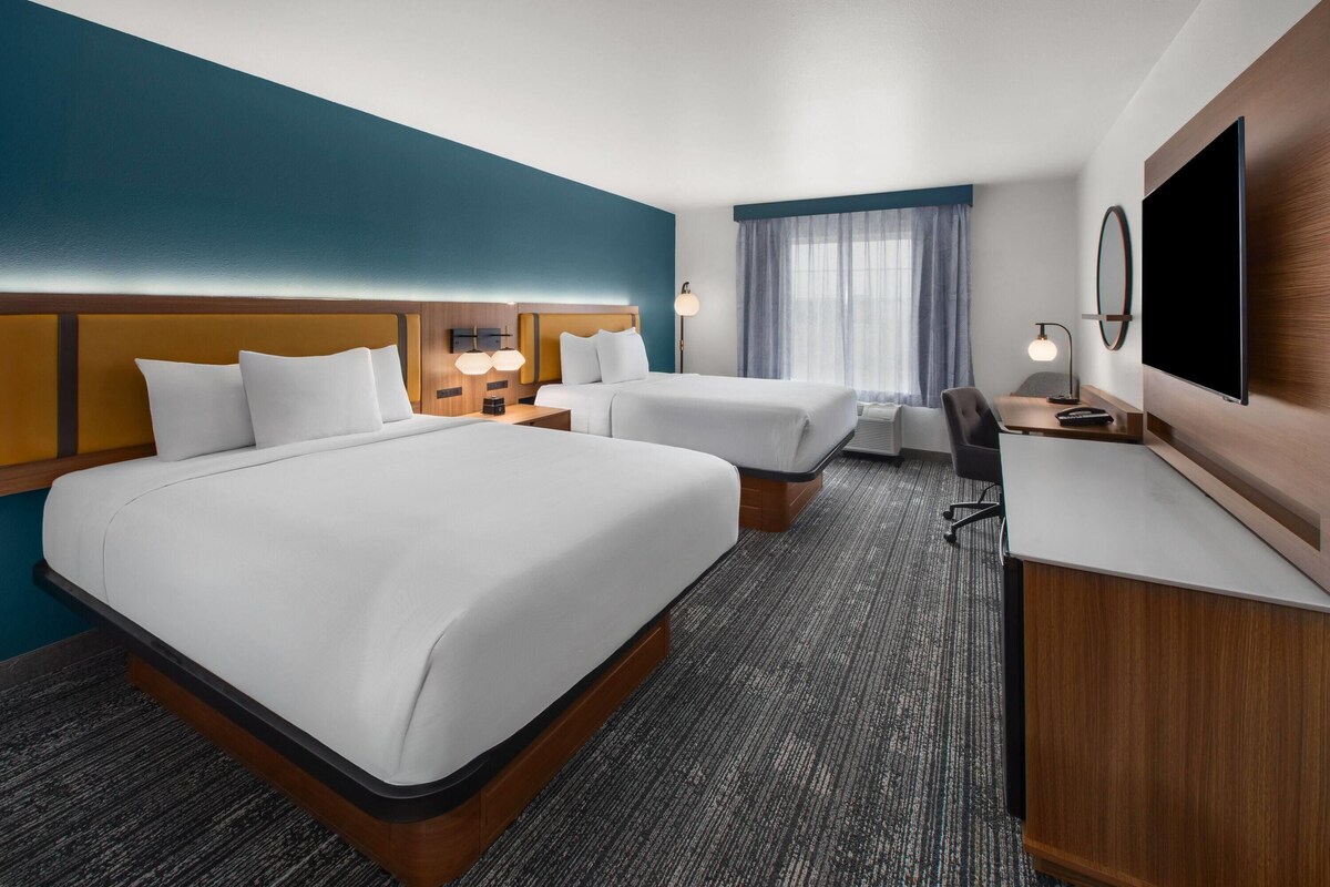 A hotel room with two beds, a desk, a TV, and a window with curtains.