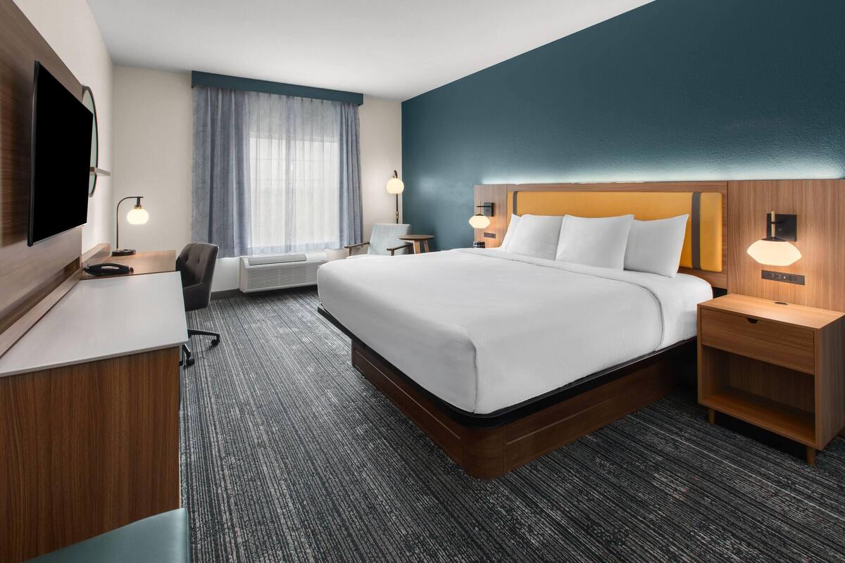 Habitación de hotel con una cama grande, un escritorio con televisión de pantalla plana, una silla, una lámpara de escritorio, una mesita de noche y una ventana con cortinas.