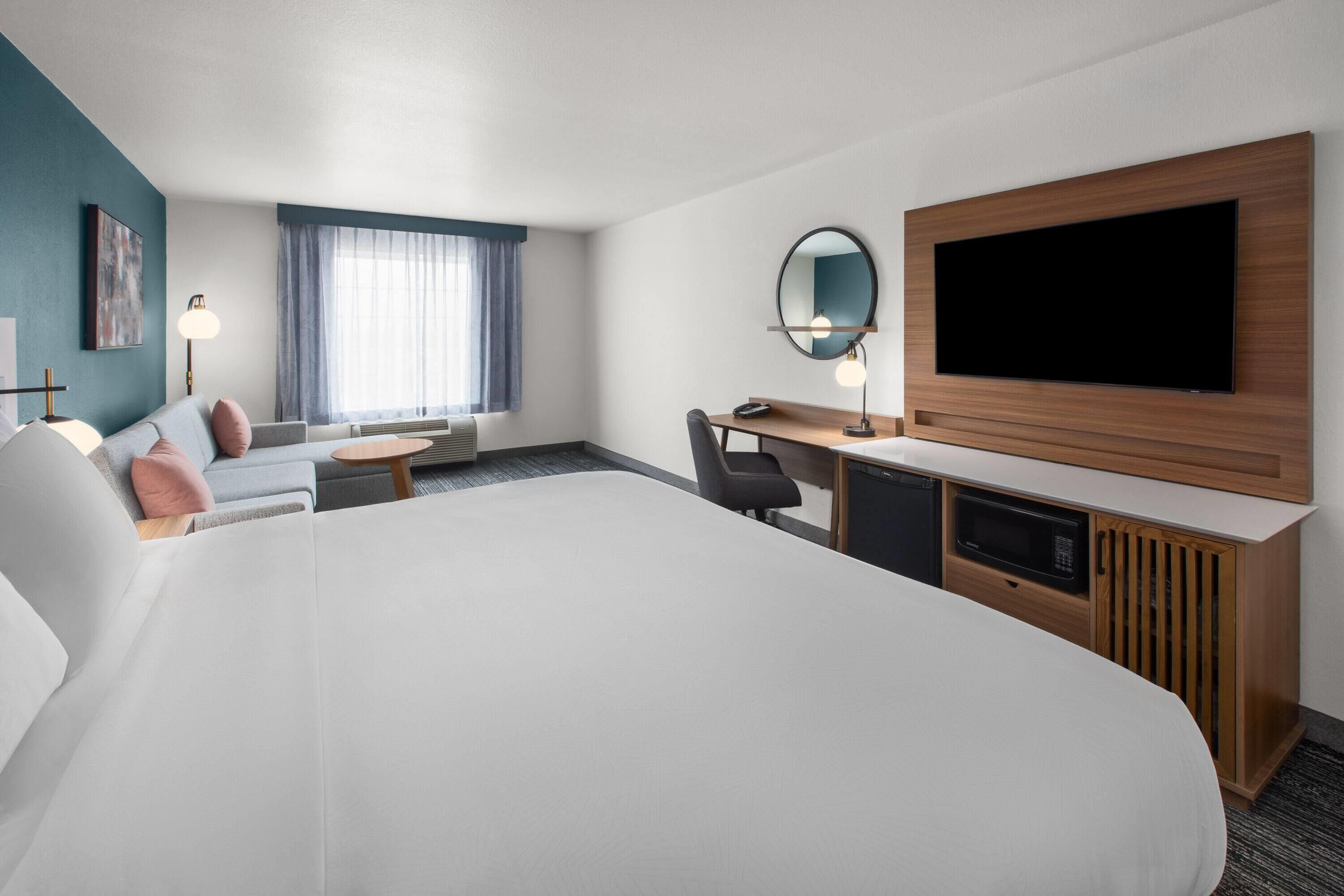 A hotel room with a large bed, a desk with a computer, a TV, a small table, and a seating area with a couch and chairs.