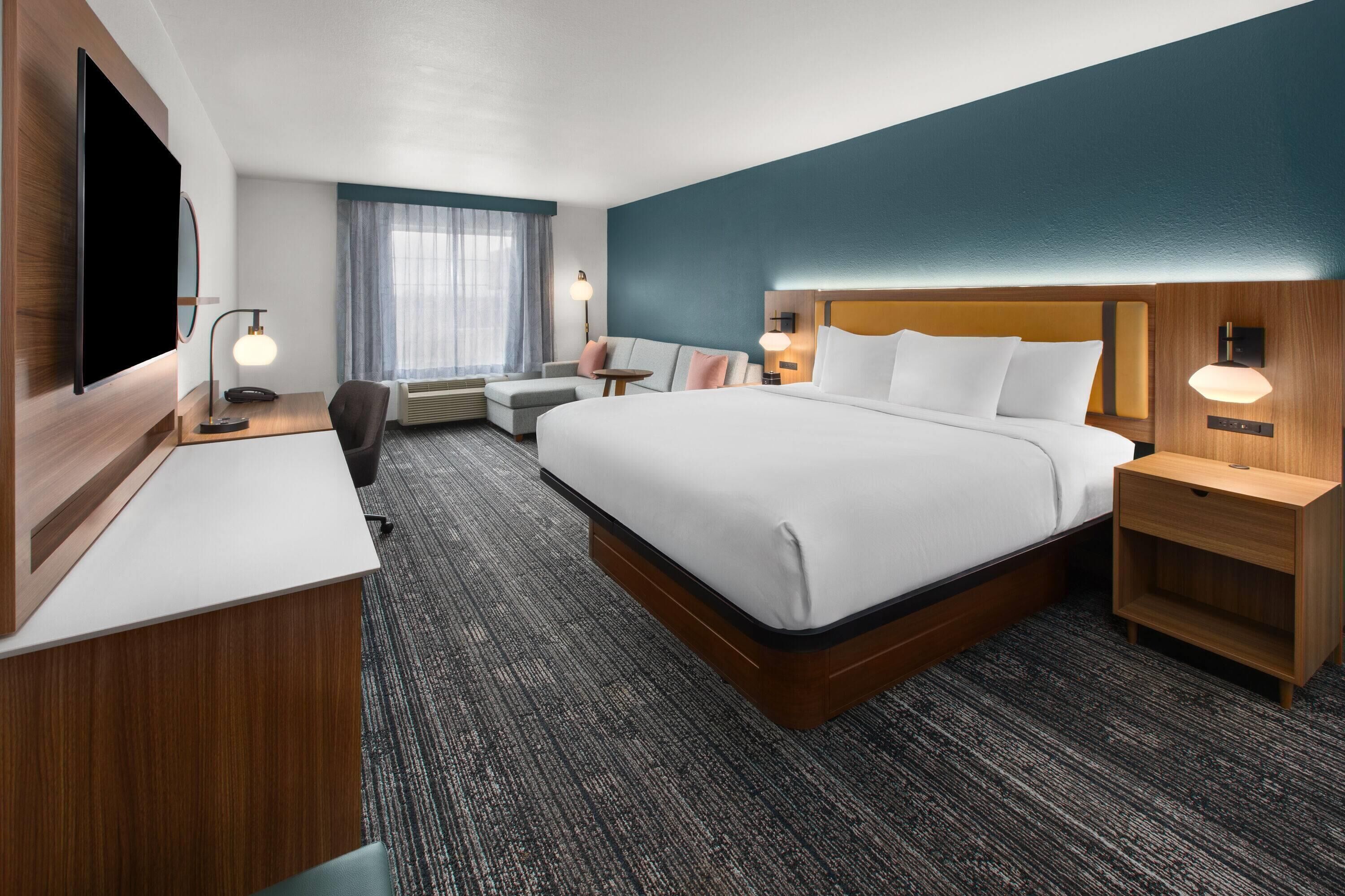 A hotel room with a large bed, a desk, a chair, a TV, and a window with curtains.
