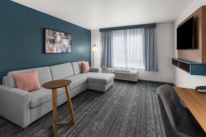 A modern living room with a sectional sofa, a round wooden side table, a flat-screen TV mounted on the wall, and a large window with sheer curtains. - La Quinta Inn & Suites by Wyndham Minot (Minot)