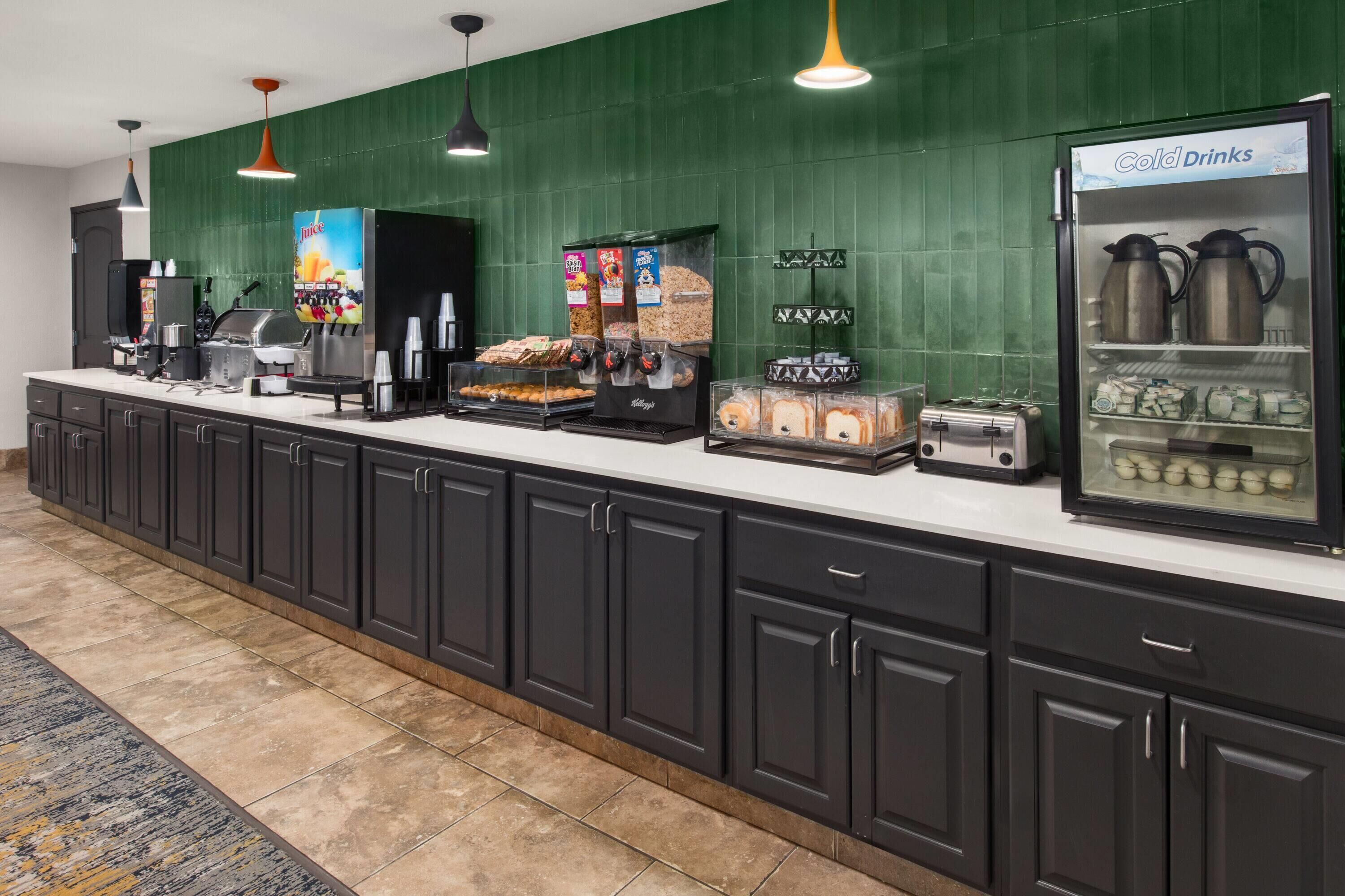 Un espace dédié au petit-déjeuner dans l’hôtel, avec un long comptoir, des machines à café, un distributeur de jus de fruits et un réfrigérateur rempli de boissons fraîches.