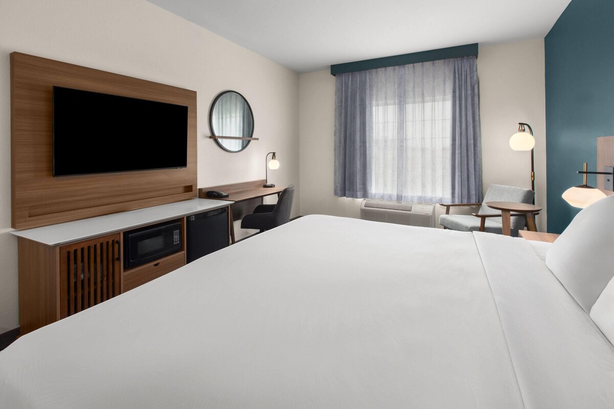 A hotel room with a large bed, a desk with a chair, a television, a mirror, a window with curtains, and a lamp.