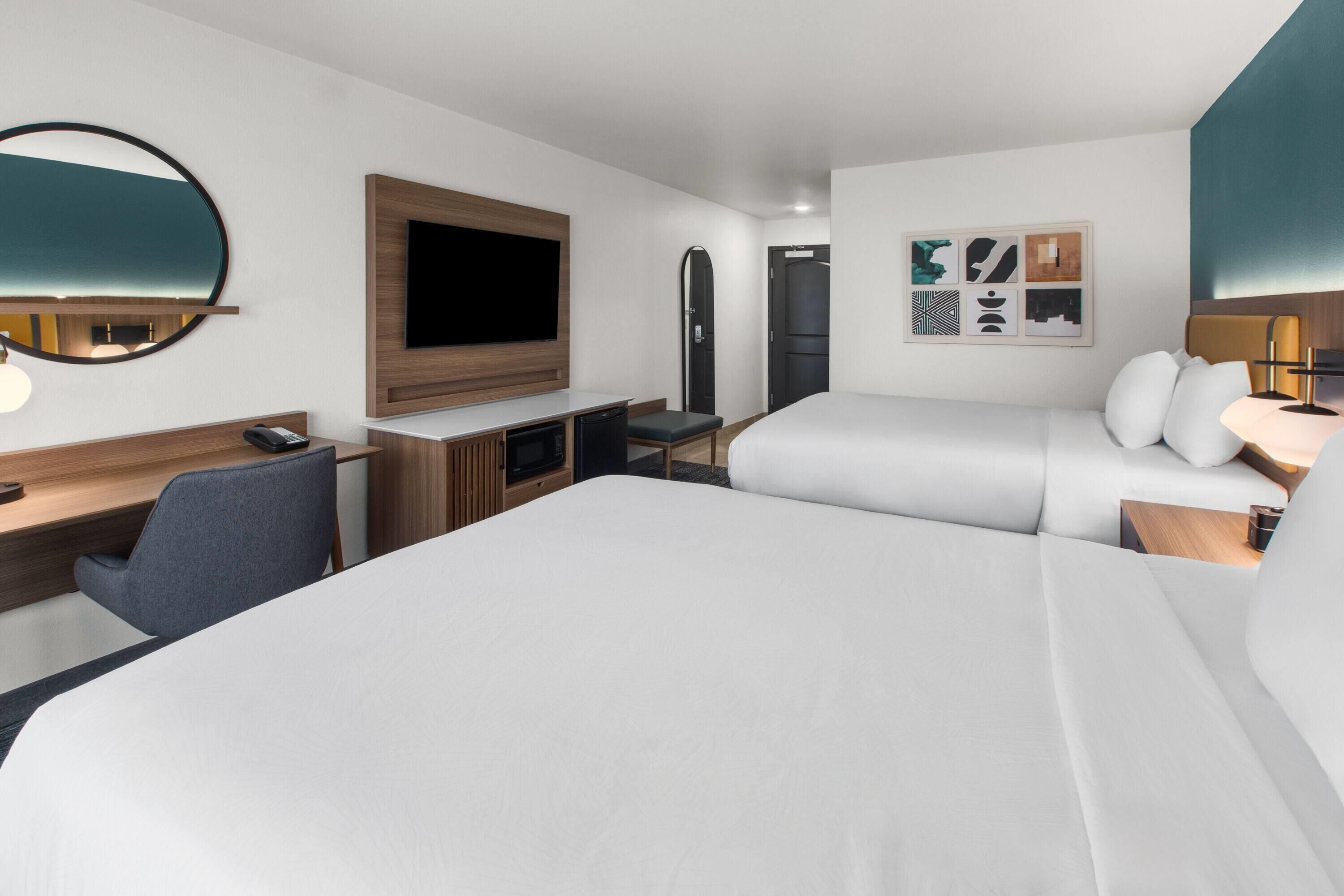 A hotel room with two beds, a desk, a TV, and artwork on the wall.