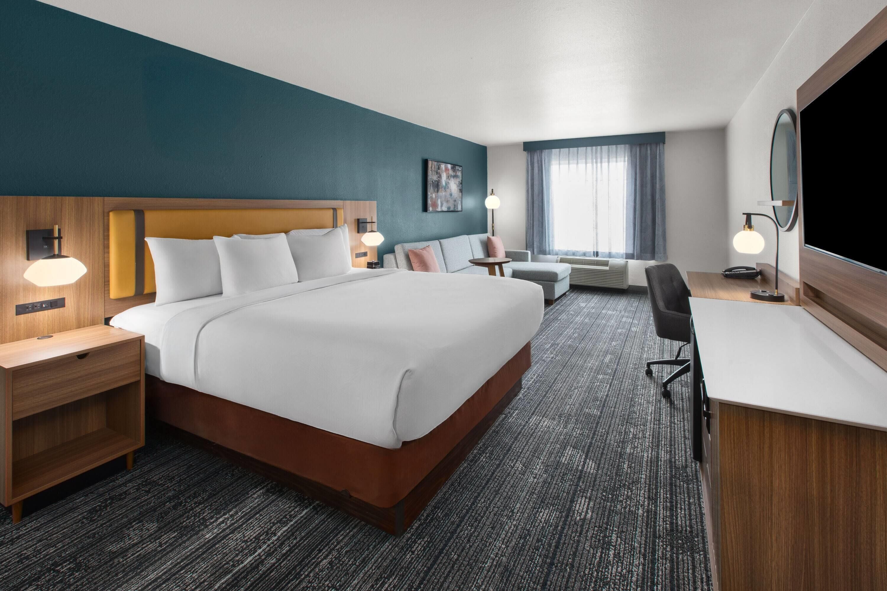 Une chambre d’hôtel avec un grand lit, un bureau, une chaise, une table de chevet, une lampe et une fenêtre avec des rideaux.