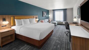 Une chambre d’hôtel avec un grand lit, un bureau, une chaise, une table de chevet, une lampe et une fenêtre avec des rideaux.