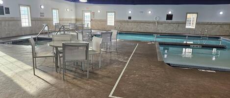 An indoor swimming pool area with multiple lanes, a lounge area with chairs and tables, and a tiled floor.