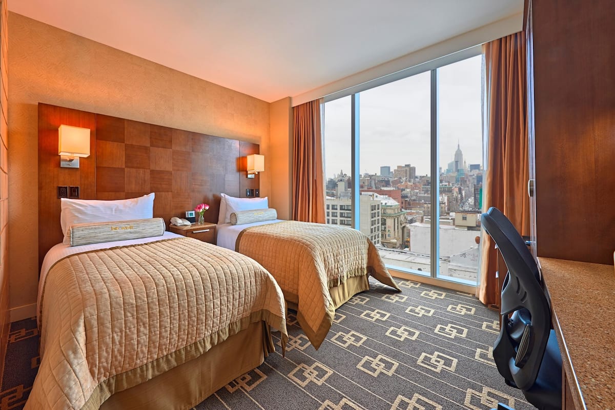 A hotel room with two beds, a large window with a city view, and a desk with a chair.
