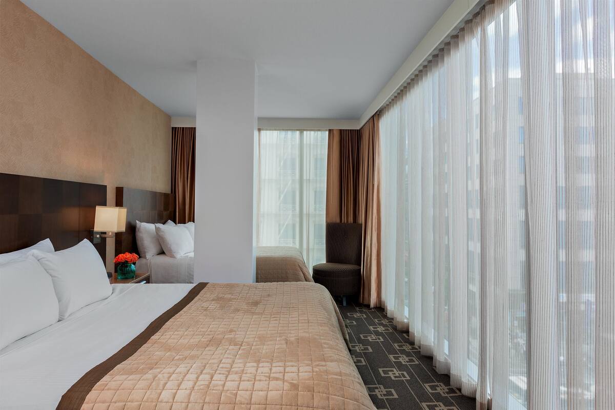 A hotel room with a large bed, a sitting area with a chair, a small table with a vase of flowers, and a view through sheer curtains.