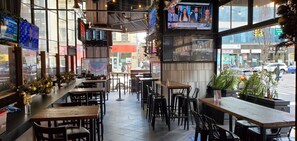Un restaurante con varias pantallas de televisión mostrando noticieros, diferentes opciones de asientos que incluyen mesas y sillas, y vista a la calle.