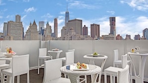 Una terraza en la azotea con mobiliario de mimbre blanco, vistas al horizonte de la ciudad y un plato de comida.