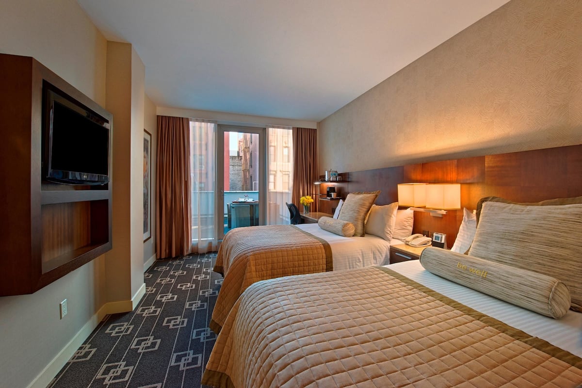 A hotel room with two beds, a TV, a desk with a lamp, and a view of the city through large windows.