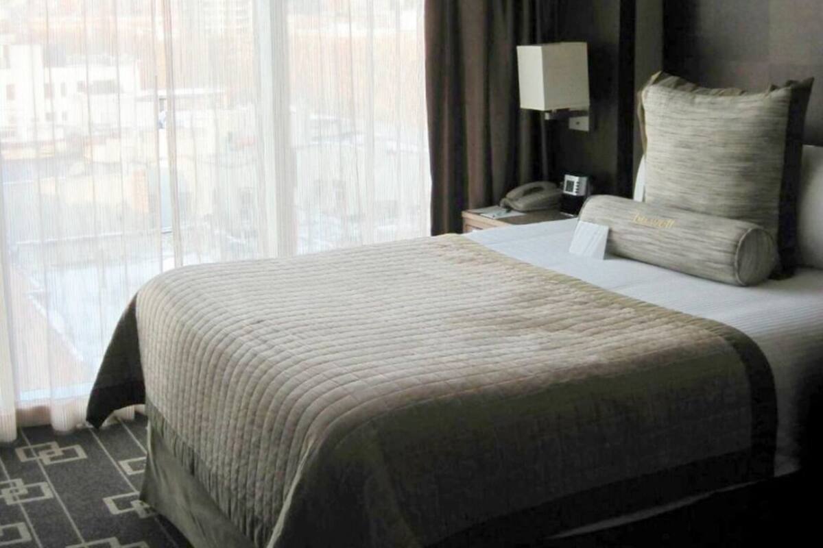 A hotel room with a large bed, a nightstand, a lamp, and a view of the city through sheer curtains.