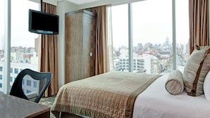 Una habitación de hotel con una cama grande, un televisor montado en la pared, un escritorio y una silla. La habitación tiene ventanas amplias con vistas a la ciudad.