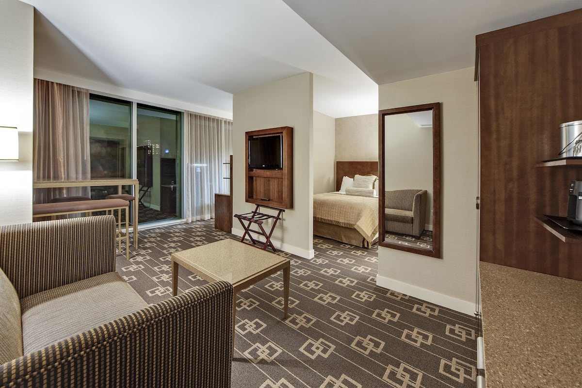 A hotel room with a sofa, a small table, a bed with a headboard, a television mounted on the wall, and a large mirror.